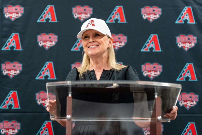 Person wearing a white Arizona Diamondbacks cap and black top stands behind a clear podium with a backdrop featuring repeated Arizona Diamondbacks logos and text.