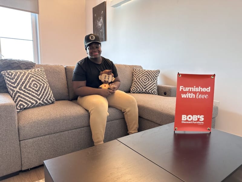 Person sitting on a beige fabric sectional sofa with patterned throw pillows, holding a plush toy, next to a red sign reading "Furnished with love BOB'S Discount Furniture."