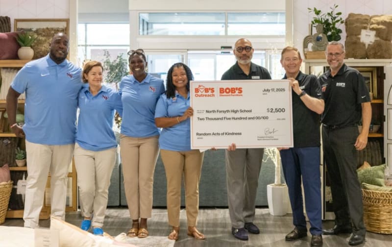 Group of seven people holding a large check from Bob's Discount Furniture dated July 17, 2025, for $2,500 to North Forsyth High School for Random Acts of Kindness. Four individuals wear light blue shirts with "NF" logo, and three wear black Bob's Discount Furniture shirts. The setting is an indoor space with shelves and plants.