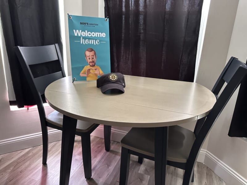 Welcome home banner from Bob's Furniture with a wooden dining table and two black chairs, featuring a black Boston Bruins cap on the table.
