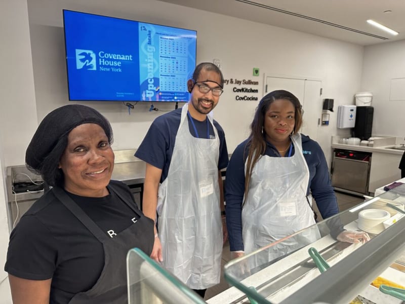 Three people wearing aprons and gloves stand behind a food service counter at Covenant House New York's CovKitchen, with a digital screen displaying intake schedules for October to December 2023 in the background.