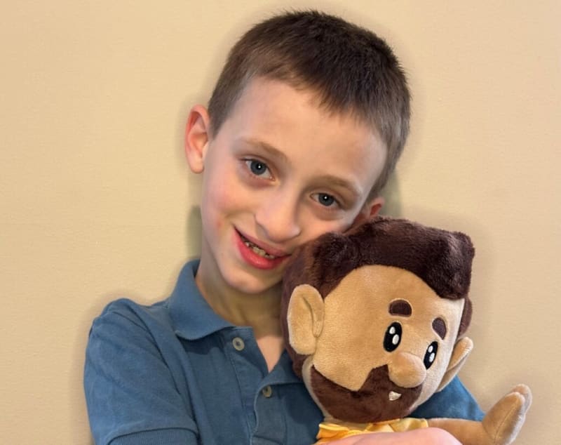 Young boy wearing a blue collared shirt holding a plush toy with brown hair, beard, and big eyes against a beige wall background.