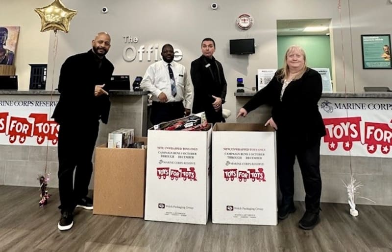 Four people stand behind two large white donation boxes labeled "Toys for Tots" with text about new, unwrapped toys for a Marine Corps Reserve campaign, in an office setting.