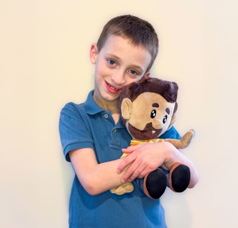 Young boy wearing a blue polo shirt hugging a plush doll with brown hair, beard, and yellow bow tie against a plain light background.