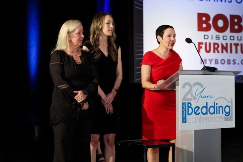 Three women stand on stage at the 20 Years Furniture Today Bedding Conference, with one speaking at a podium displaying the event logo.