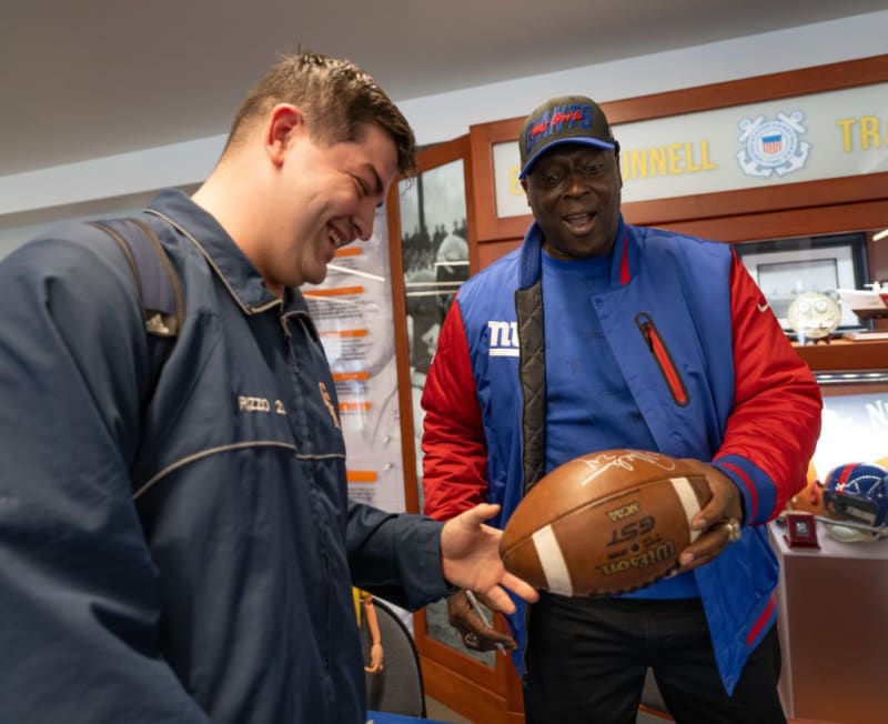Two men indoors, one wearing a blue and red New York Giants jacket and cap holding a Wilson GST NFL football, the other in a dark blue jacket reaching toward the ball.