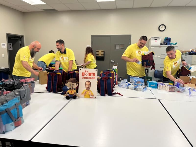 Volunteers wearing yellow shirts packing navy blue and red backpacks with supplies like Swiss Miss hot cocoa in a community outreach event labeled "BOB'S Outreach."