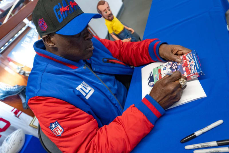 Man wearing a red and blue New York Giants NFL jacket and black cap signing a sports card on a blue table with Sharpie markers nearby.