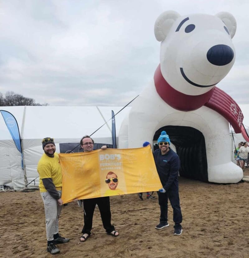 Three people stand on a sandy surface holding a yellow banner that reads "BOB'S FURNITURE" in front of a large inflatable white bear wearing a red scarf, likely at an outdoor event or promotion.