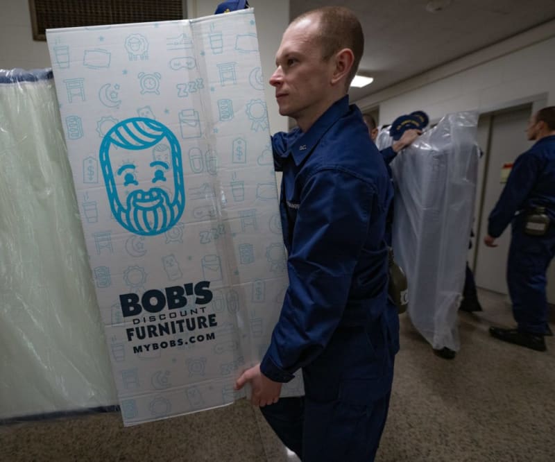 Person in navy uniform carrying a boxed mattress from Bob's Discount Furniture with logo and website mybobs.com visible, indoors.