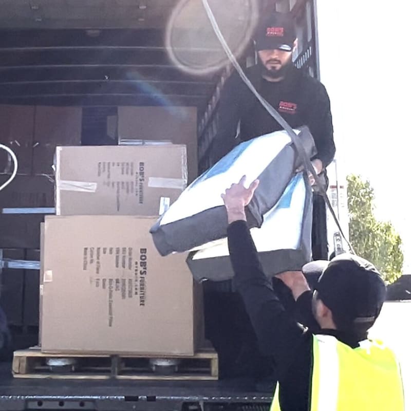 Two workers unloading large gray and white cushions from a truck filled with cardboard boxes labeled "BOB'S Furniture," one wearing a yellow safety vest and black cap.
