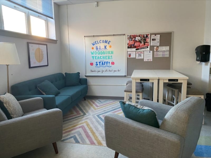 Welcome sign in a cozy lounge area with teal and gray upholstered sofas, a patterned rug, white tables, and a bulletin board. The sign reads: "WELCOME BACK WOODBURN TEACHERS and STAFF! thanks you for all that you do!"