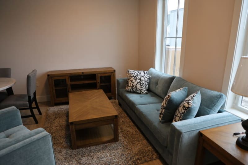 Modern living room with a teal fabric sofa featuring patterned throw pillows, wooden coffee table, side table with lamp, and a textured area rug.