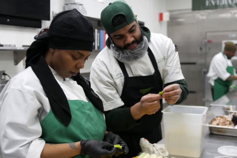 Two chefs wearing white shirts, green aprons, and black gloves prepare food in a commercial kitchen with stainless steel counters and containers.