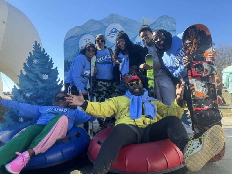 Group of people wearing blue "PLUNGE" sweatshirts posing outdoors with snow tubes, skis, and a snowboard in front of a winter-themed backdrop.