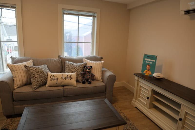 Living room with a gray fabric sofa featuring patterned and striped throw pillows, including one with "Stay Awhile" text, a wooden coffee table, and a cream-colored media console with a "Welcome Home" sign and football on top.
