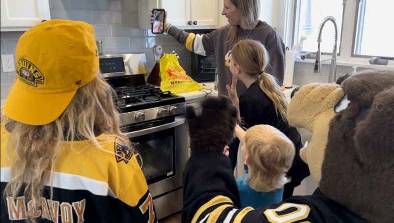 A group of children and a person in a Boston Bruins mascot costume gather in a kitchen, with one child wearing a yellow Bruins cap and jersey, and another taking a selfie on a smartphone.