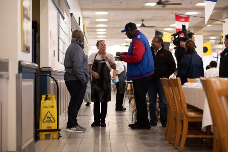 A group of people standing and talking in a hallway of a restaurant or cafeteria, with one person wearing a striped apron and others in casual clothing. A yellow caution wet floor sign is visible on the left. Chairs and tables with white tablecloths are on the right.