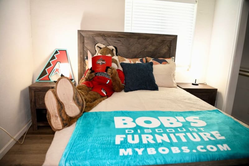 A person in a brown animal mascot costume wearing a red jersey reclines on a beige bed with a wooden headboard, reading a red booklet. A turquoise blanket at the foot of the bed displays white text: "BOB'S DISCOUNT FURNITURE MYBOBS.COM." The room features two wooden nightstands, one with a lamp shaped like a stylized letter "A."