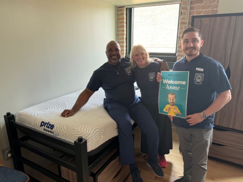 Three people wearing matching navy blue shirts stand around a black metal-framed bed with a white Prize Sleep mattress. One person holds a sign that reads "Welcome home" with a small image of a child in a yellow shirt. The scene suggests a home or dorm room setup, highlighting the Prize Sleep mattress brand.