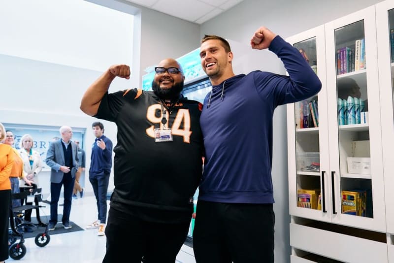 Two men flexing their arms in a bright indoor setting, one wearing a black sports jersey with number 94 and a lanyard, the other in a navy blue hoodie, standing near white cabinets with books and products.