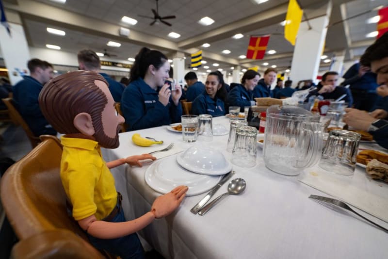 A group of people in blue uniforms seated around a long table with white tablecloth, glass cups, a water pitcher, utensils, and a banana, in a large indoor dining area decorated with hanging flags.