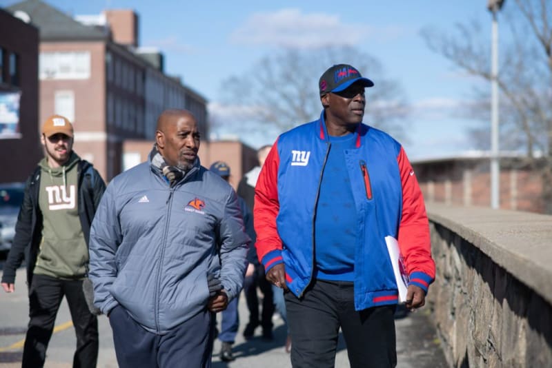Three men walking outdoors on a sunny day, wearing casual jackets and hoodies. One man wears a blue and red New York Giants jacket and cap, another wears a gray Adidas jacket with a Coast Guard emblem, and the third wears a green hoodie with a white "ny" logo and a tan cap. The background shows a brick building and a stone wall along the sidewalk.