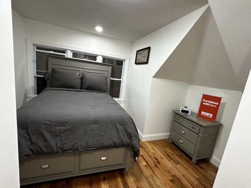 Bedroom with gray wooden bed frame and matching nightstand on hardwood floor, featuring dark gray bedding and a sign reading "Furnished with love BOB'S."