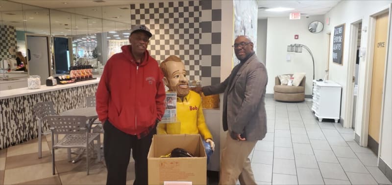 Two men stand indoors near a mannequin wearing a yellow "Bob's" shirt, with a large cardboard box in front. The setting includes tiled floors, a checkered wall, and office doors labeled "OFFICE."