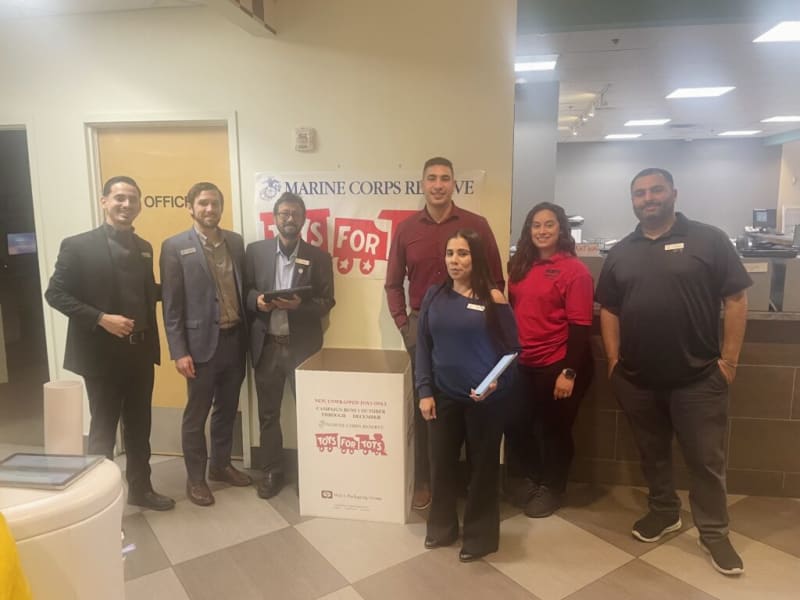 Group of six people standing indoors near a large white donation box labeled "Toys for Tots" with a Marine Corps Reserve banner and text: "NEW, UNWRAPPED TOYS ONLY! CAMPAIGN RUNS 1 OCTOBER THROUGH 1 DECEMBER (MARINE CORPS RESERVE)" in the background.