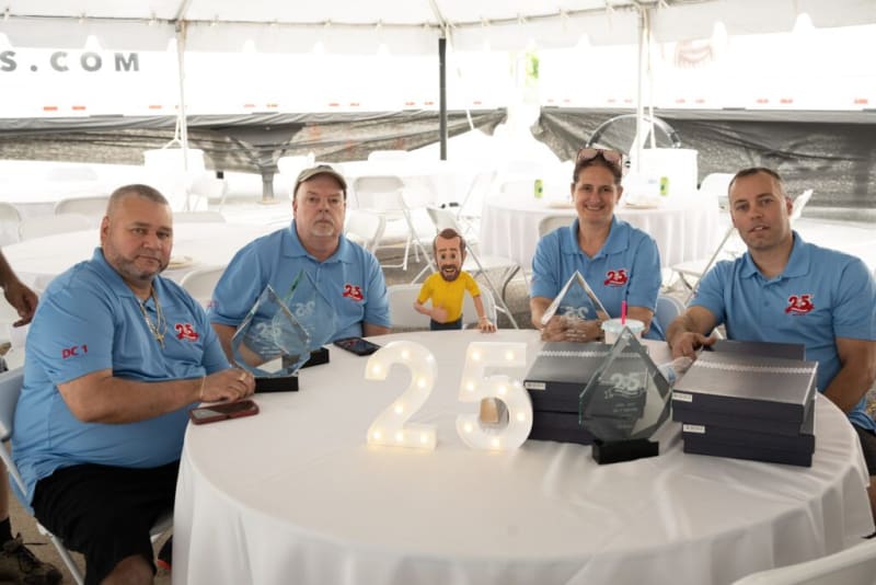 Four men in blue polo shirts with a "25" logo sit around a round table with illuminated numbers "25," holding glass awards and surrounded by black boxes, celebrating a 25th anniversary event.