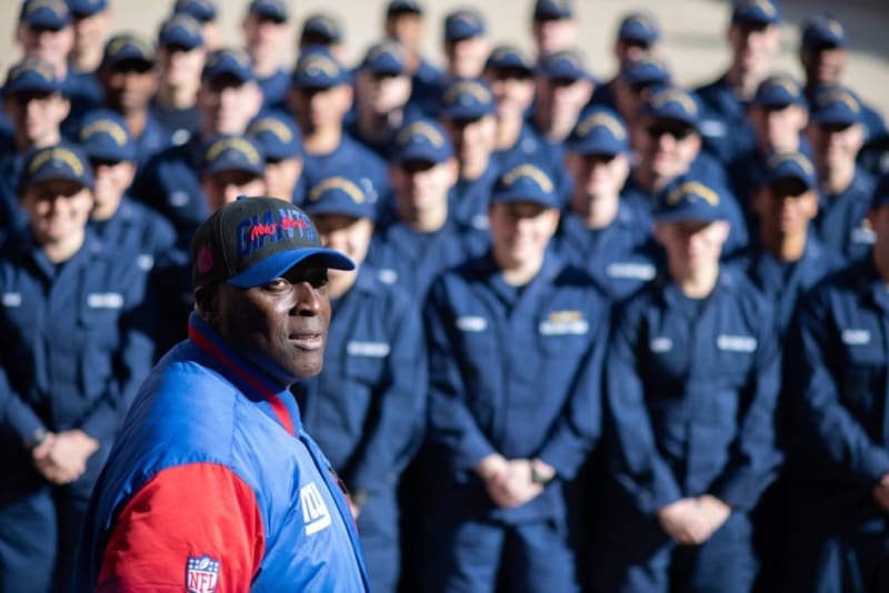 A group of uniformed personnel in navy blue standing in formation behind a person wearing a blue and red NFL jacket and a colorful cap.