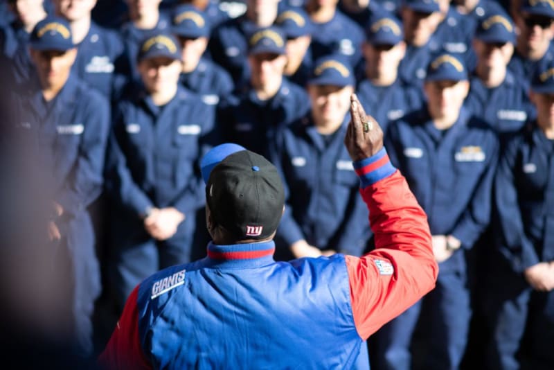 Man wearing a New York Giants jacket and cap addressing a group of people in navy blue uniforms, likely a team briefing or training session.