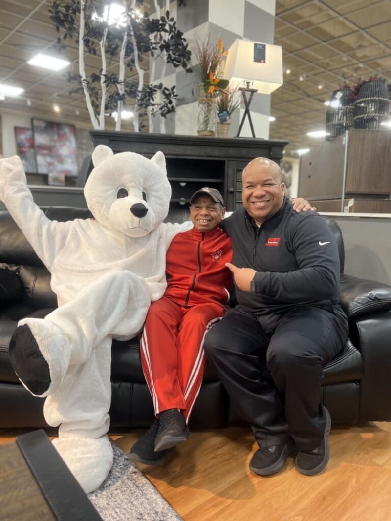 Three people sitting on a black leather couch in a furniture store; one person in a white bear costume, one in a red tracksuit with white stripes, and one in a black Nike jacket and pants.