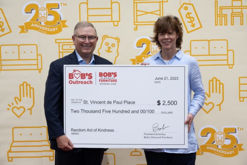 Two people holding a large check from Bob's Discount Furniture dated June 21, 2023, for $2,500 payable to St. Vincent de Paul Place, memo: Random Act of Kindness. Background features yellow illustrations of furniture and the 25th anniversary logo.