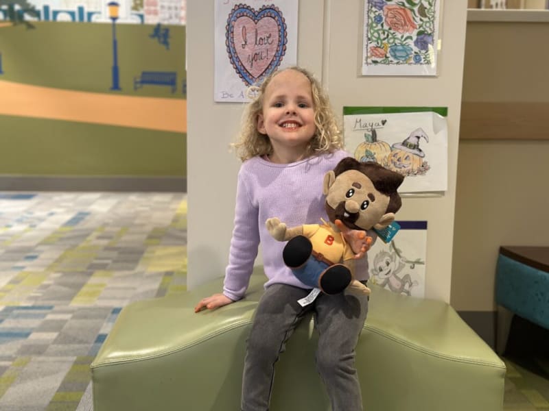 Child with curly blonde hair wearing a lavender sweater and gray pants sits on a green padded bench holding a plush doll with brown hair, mustache, yellow shirt, and blue pants. Behind are colorful drawings including a heart with "I love you!" and pumpkins labeled "Maya." The setting appears to be a children's play or waiting area with patterned carpet and soft seating.