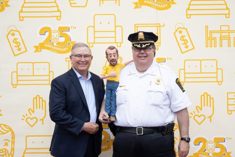 Two men standing in front of a backdrop with yellow furniture icons and "25th Anniversary" text, one in a police uniform and the other in a dark suit.