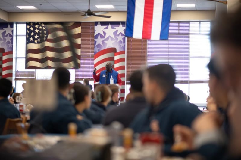 Man speaking at a podium in a room decorated with American flag-themed banners and a hanging red, white, and blue flag, addressing an audience seated at tables.