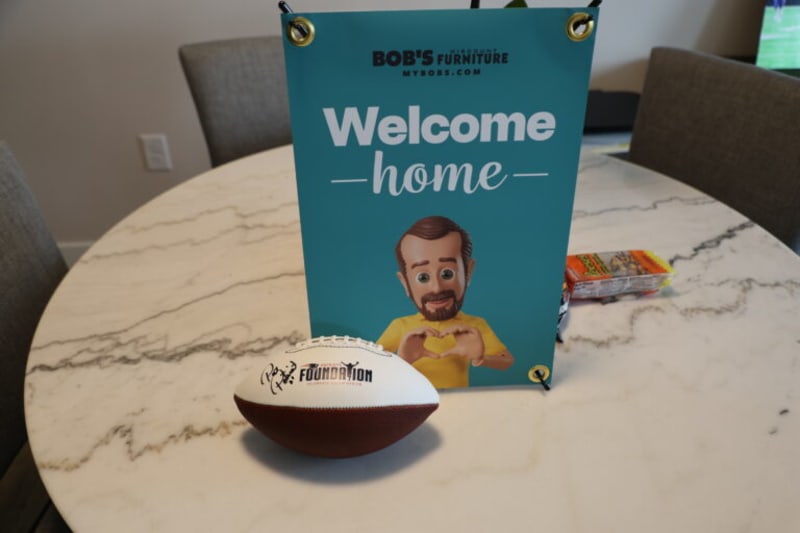 Blue welcome banner from Bob's Discount Furniture with text "Welcome home" and a small football with "Foundation" branding on a round white marble table.