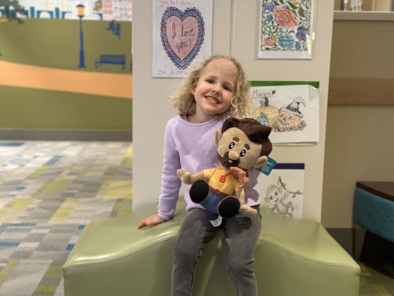 A young child with curly blonde hair wearing a lavender sweater and gray pants sits on a green cushioned bench holding a plush doll with brown hair, a beard, and a yellow shirt. Behind them are colorful drawings on the wall, including a heart with the text "you got this... I love you! Be A Star," a floral design, pumpkins with the name "Maya," and a monkey sketch. The setting appears to be a cheerful, child-friendly indoor space with patterned carpet flooring.