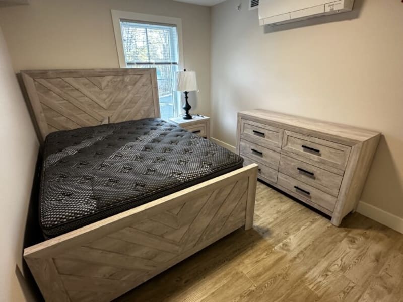 Bedroom setup with a wooden queen bed frame featuring chevron pattern, black mattress, matching light wood nightstand with lamp, and six-drawer dresser.