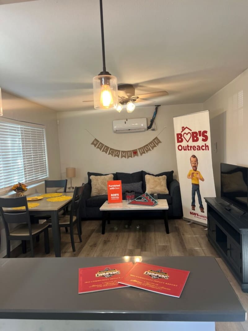 Modern living room with dark gray sofa, beige pillows, marble coffee table, and "WELCOME HOME" banner; includes Bob's Outreach signage and dining area with gray table and chairs.