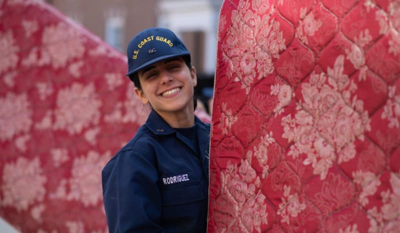 U.S. Coast Guard personnel in navy uniform and cap labeled "U.S. COAST GUARD" and name tag "RODRIGUEZ" carrying red floral-patterned mattresses.