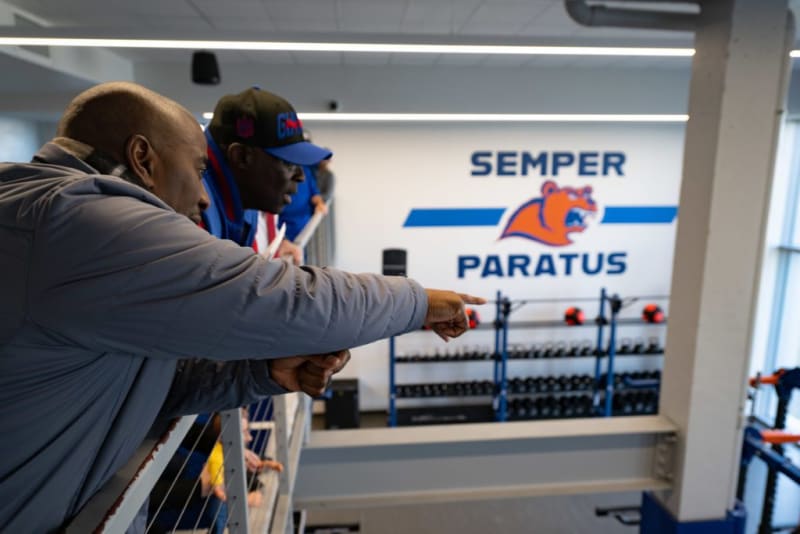 Two men leaning on a railing inside a gym, one pointing toward a wall with the text "SEMPER PARATUS" and an orange bear logo above weight racks.