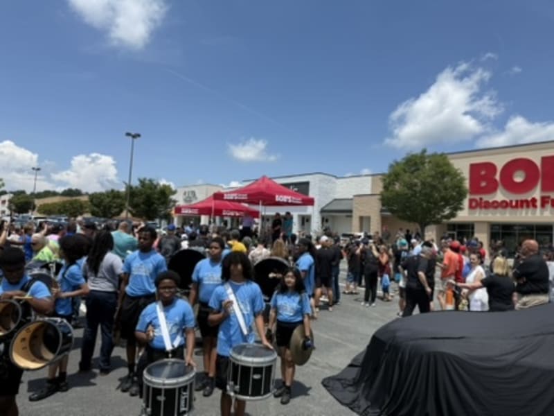 A diverse marching band in blue shirts performs with drums and cymbals in a parking lot outside a store with a red "BORD" Discount Furniture sign under a clear blue sky.