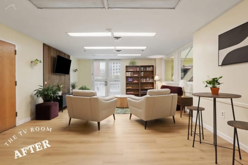 Modern TV room with beige leather armchairs, wooden flooring, wall-mounted flat-screen TV, bookshelves, and natural light from glass doors.