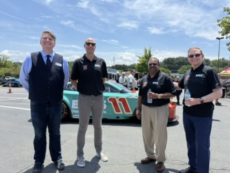 Four men stand in front of a teal and orange race car numbered 11 in a sunny parking lot, wearing business casual and branded polo shirts.