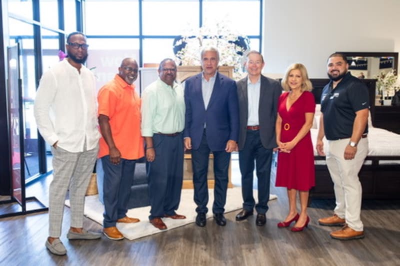 A group of six adults standing indoors on a wooden floor, dressed in business casual attire, including a red dress, suits, and casual shirts, in a well-lit modern setting with large windows and decorative elements.