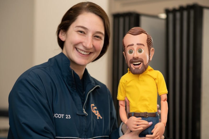Person wearing a navy blue jacket with "UGOT 23" and "CEA" logos holding a wooden puppet doll dressed in a yellow shirt and blue jeans.