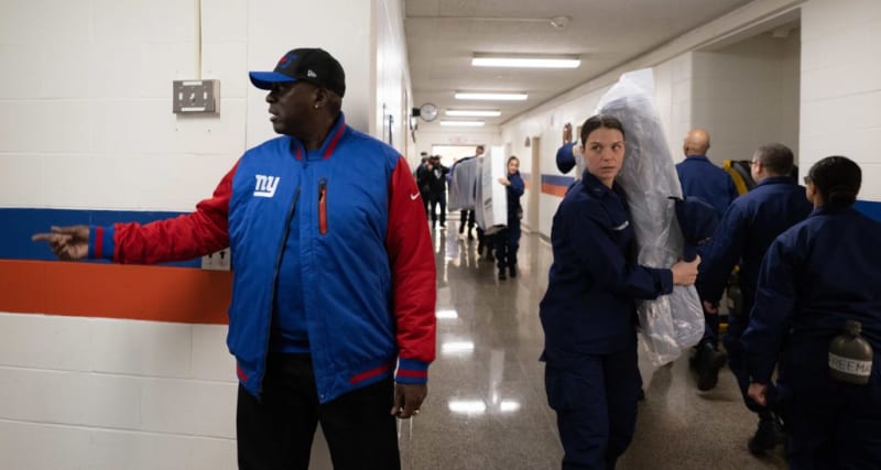 Man in blue and red New York Giants jacket points left in a hallway while uniformed personnel carry equipment and walk down the corridor.